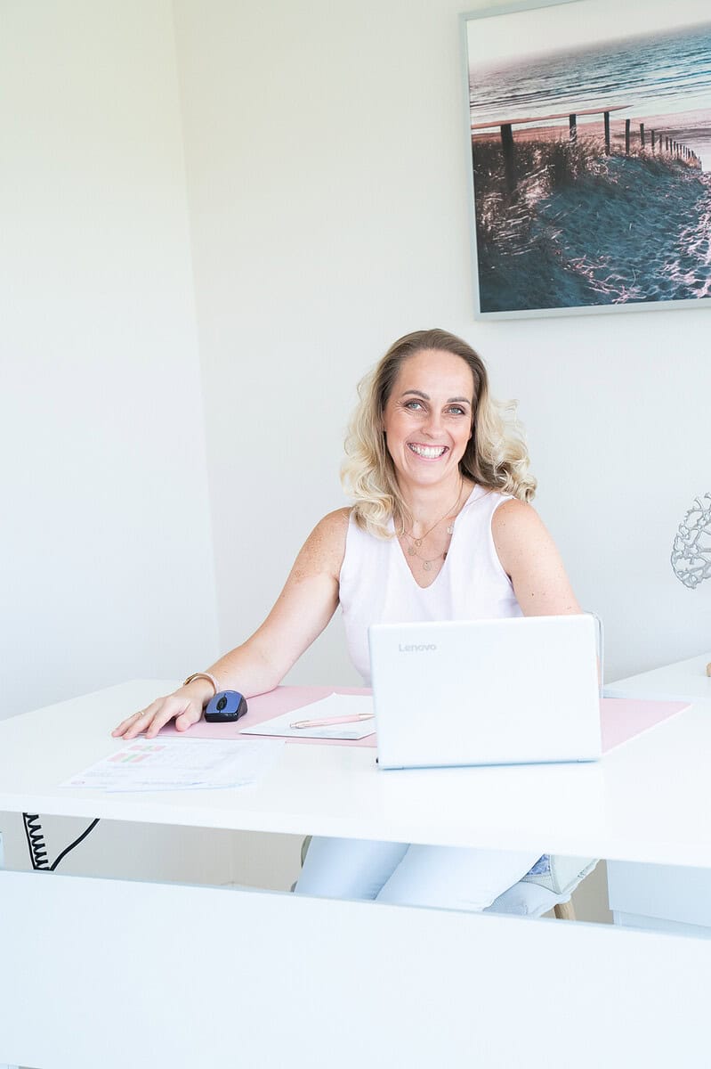 Juliane Danziger sitzt lachend an einem weißen Schreibtisch mit Laptop, Maus und Unterlagen, an der Wand hängt ein Strandbild.